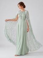 A woman models the Social Queen Beaded Printed Cape Chiffon Dress, showcasing its light green hue, intricate beaded details, patterned sheer cape sleeves, and a printed cape overlay against a plain light background.