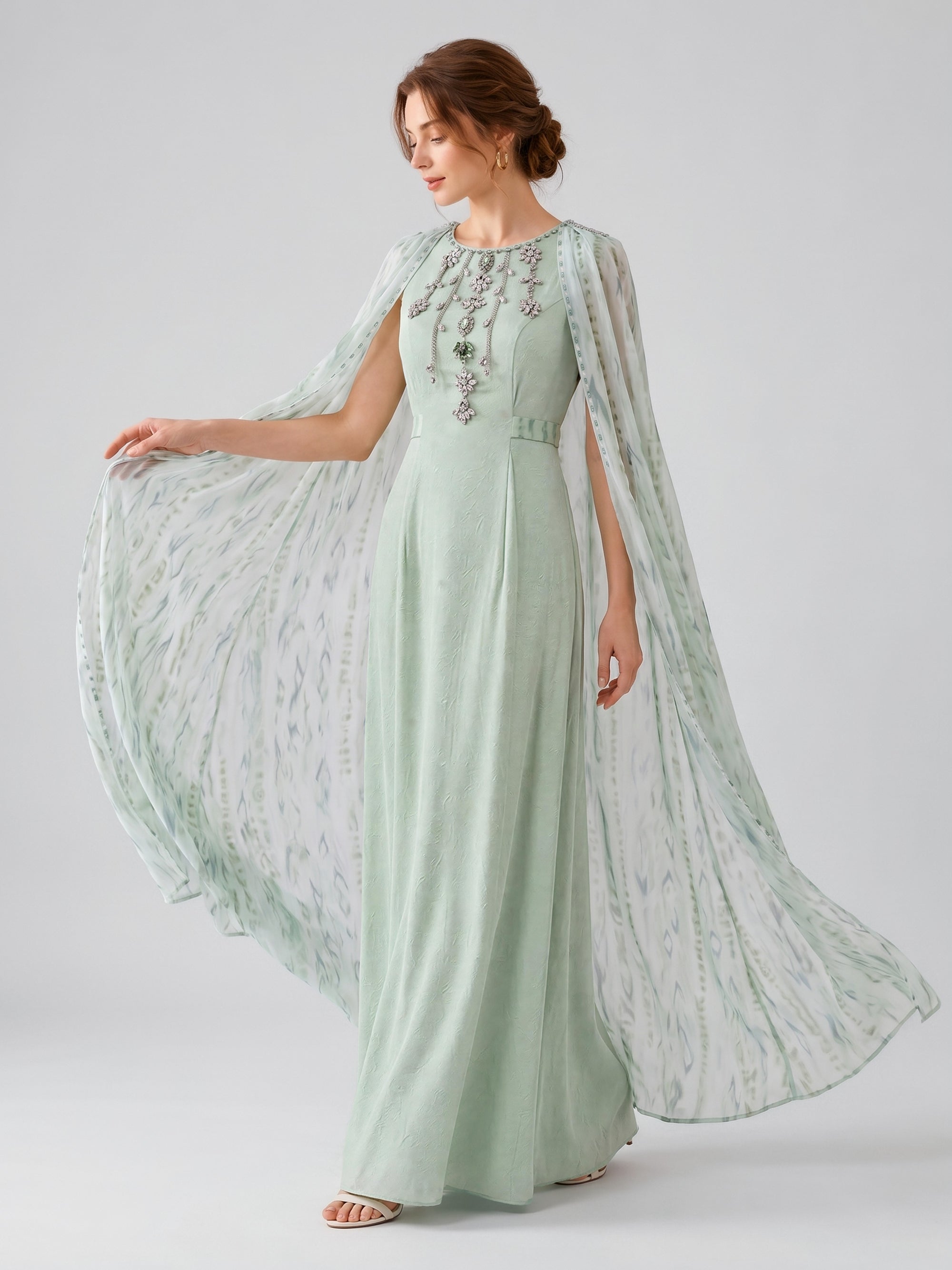 A woman models the Social Queen Beaded Printed Cape Chiffon Dress, showcasing its light green hue, intricate beaded details, patterned sheer cape sleeves, and a printed cape overlay against a plain light background.