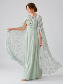 A woman models the Social Queen Beaded Printed Cape Chiffon Dress, showcasing its light green hue, intricate beaded details, patterned sheer cape sleeves, and a printed cape overlay against a plain light background.