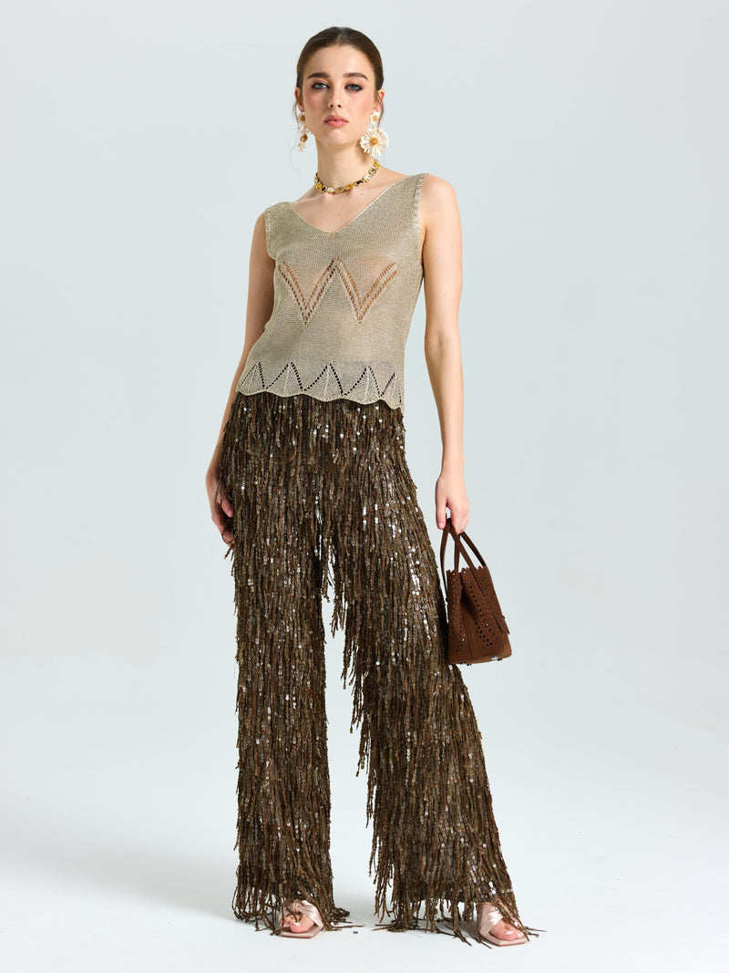A woman faces forward wearing the Social Queen Open-Knit Metallic Sleeveless Top, paired with brown fringed pants and a brown handbag, accessorized with hoop earrings and heeled sandals.