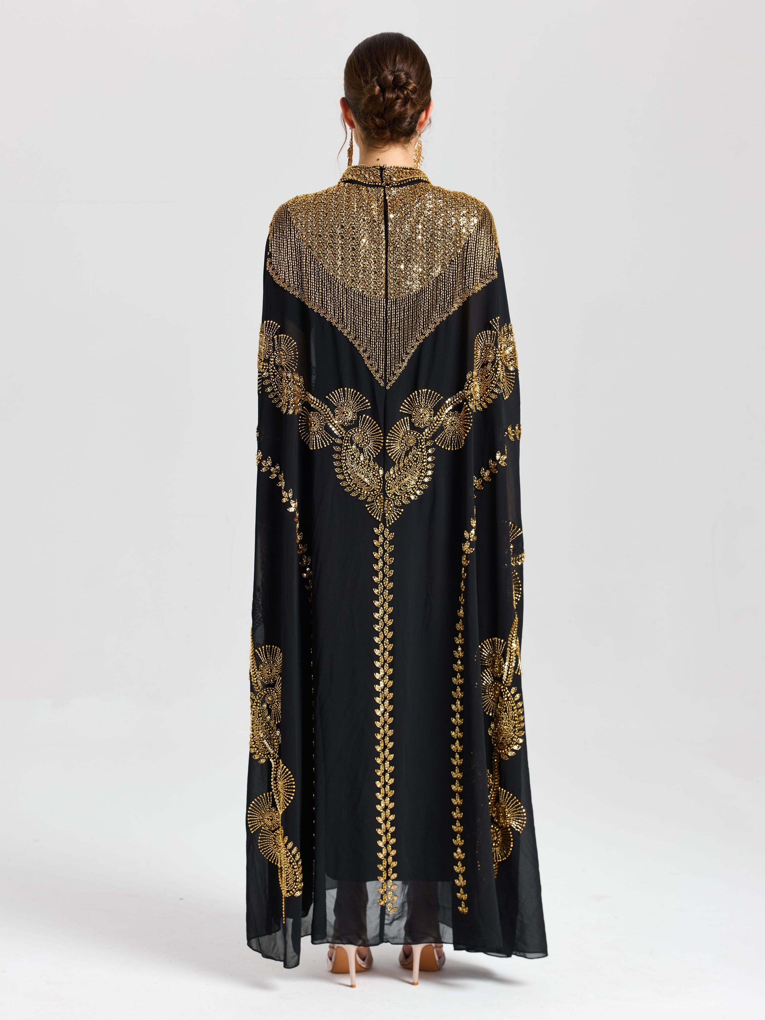 A person faces away in the Beaded Embroidered Cape Dress by Social Queen, a long black and gold chiffon piece featuring Persian-inspired embroidery—ideal for elegant evenings against a plain light background.