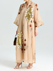 A person wears the Social Queen Placement Embroidered Linen Dress in beige with gold sequins, floral and brown leaf patterns, damask scrolls, styled with heeled sandals and a small brown handbag.