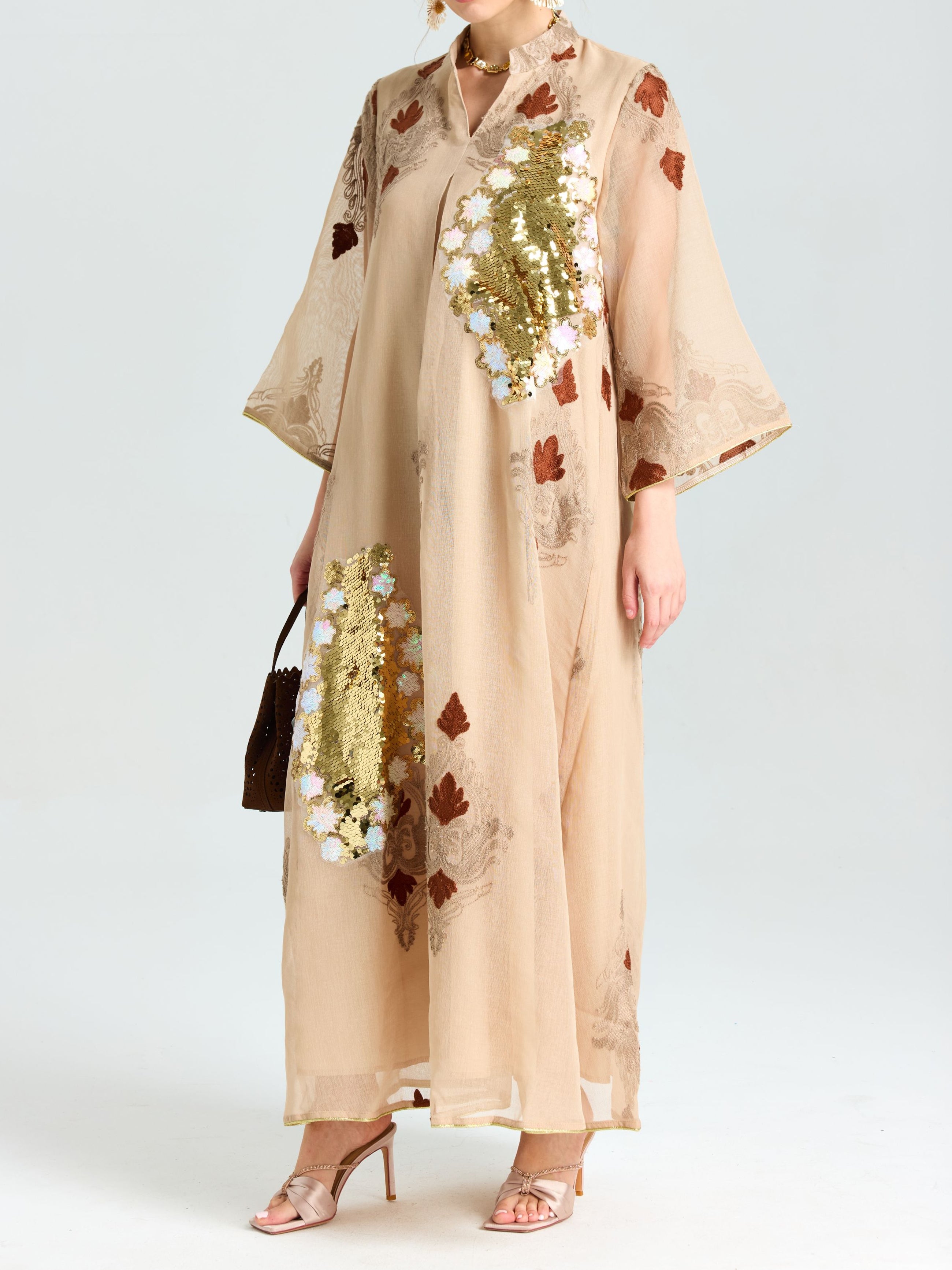 A person wears the Social Queen Placement Embroidered Linen Dress in beige with gold sequins, floral and brown leaf patterns, damask scrolls, styled with heeled sandals and a small brown handbag.