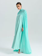 A woman in the Social Queen Beaded Cape Scarf Gown, featuring a flowing light blue chiffon cape and sequin-detailed bodice, stands against a plain light background.