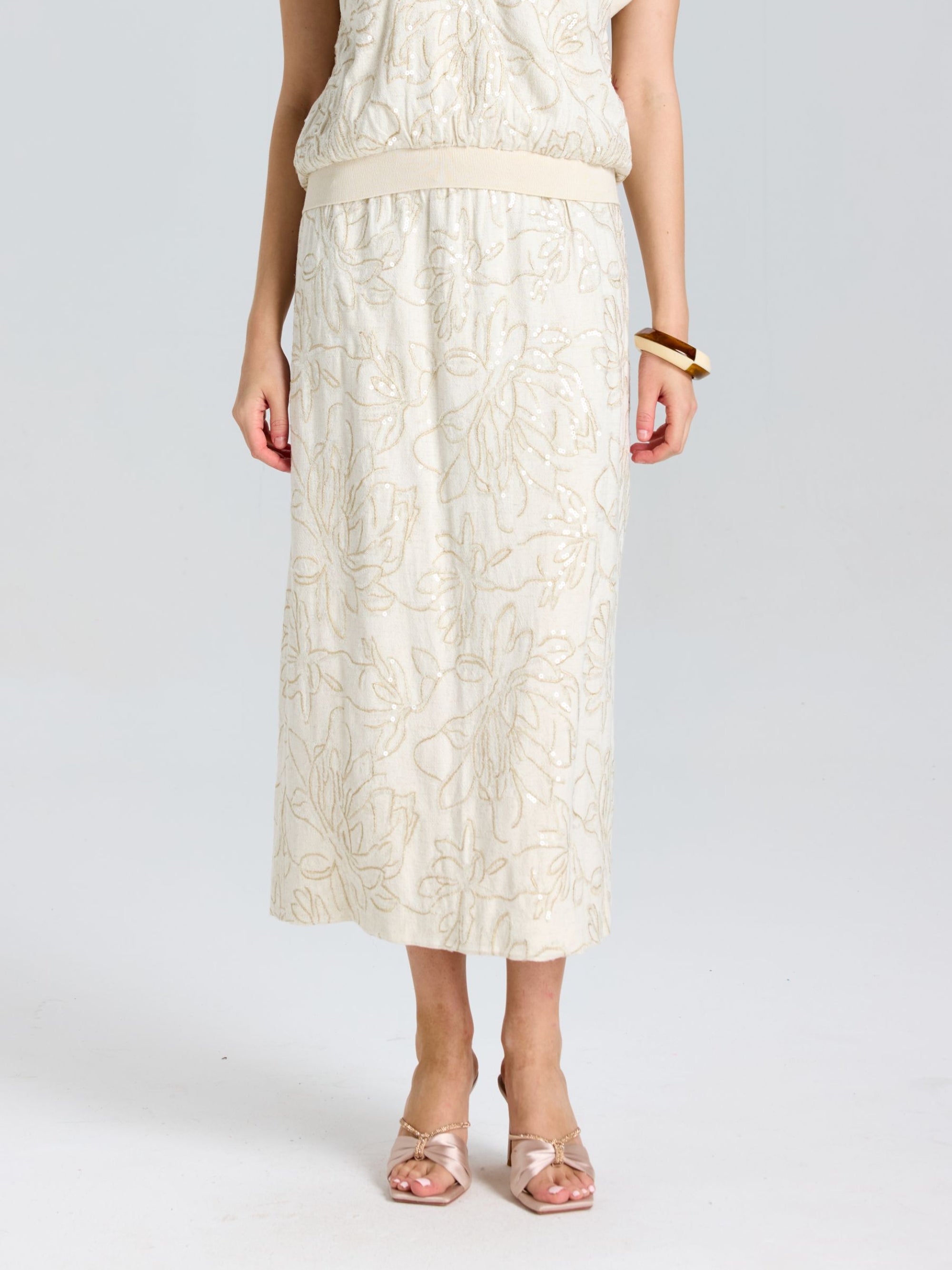 A person wearing the Social Queen Floral Embroidered Linen Midi Skirt with a light top, gold bracelet, and light pink open-toe shoes.