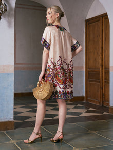 Wearing the Social Queen Paisley Garden Print Jacquard Silk Dress and gold heels, a woman stands in a tiled hallway with arched doorways and wooden doors, holding a bamboo handbag.
