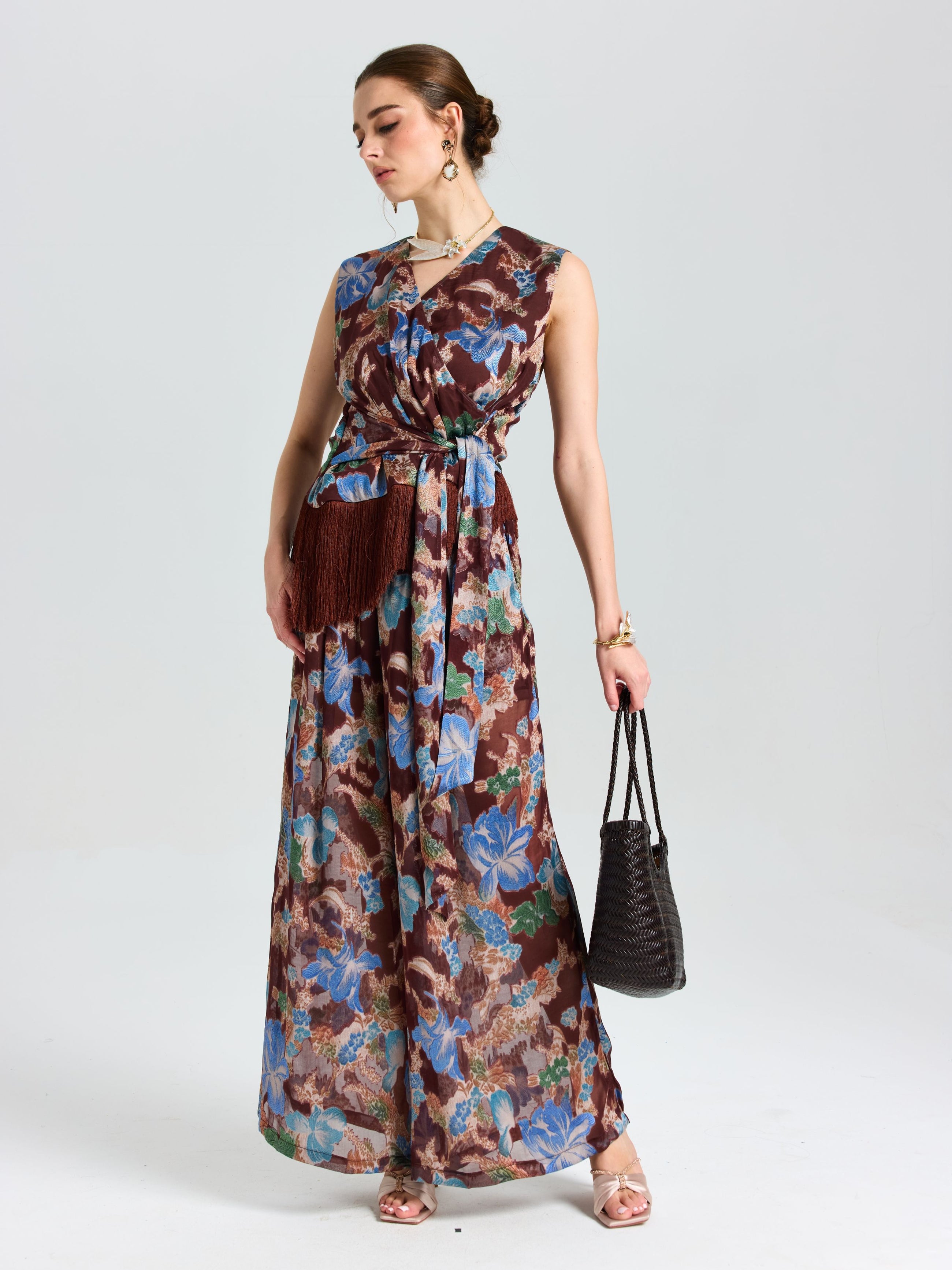 A woman models Social Queen’s Floral Print Wide-Leg Pants with a brown belt and fringe pouch, black woven handbag in hand—a relaxed, vacation-ready look set against a plain light background.