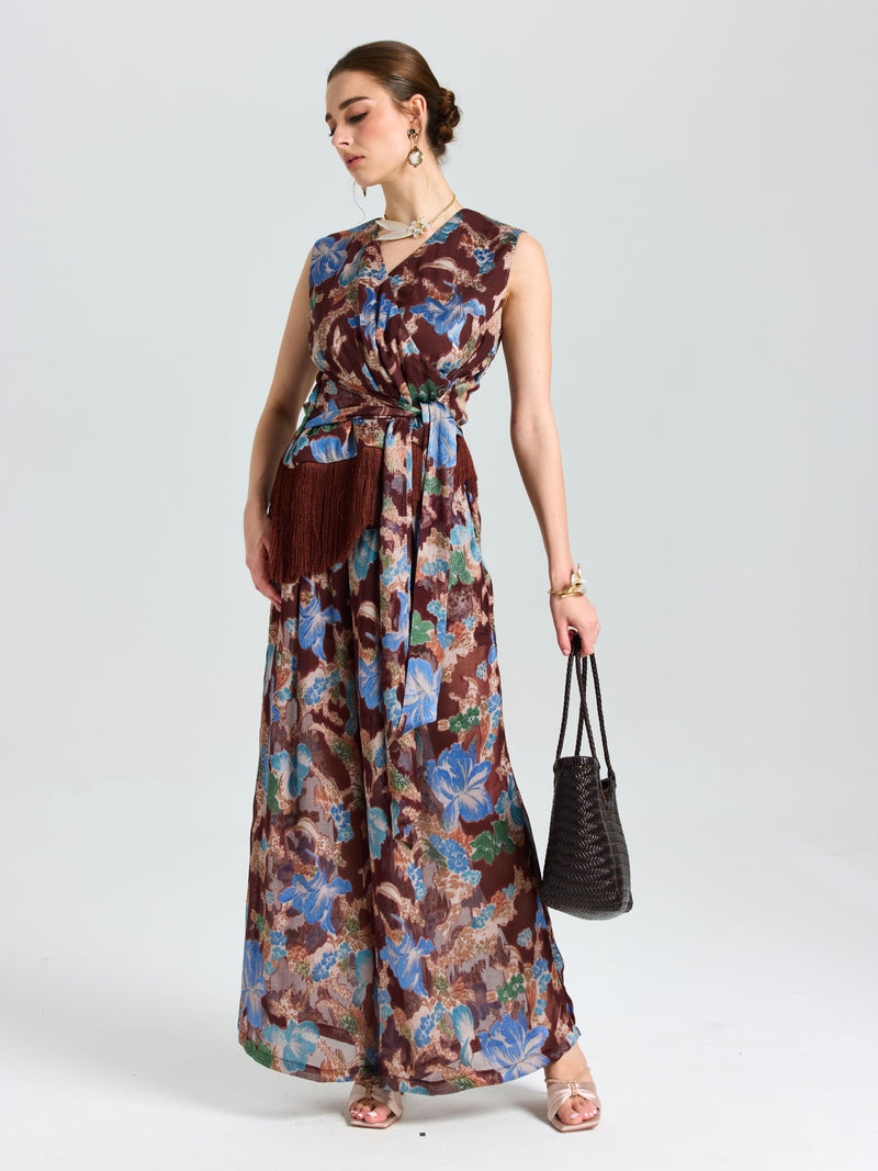 A woman models Social Queen’s Floral Print Wide-Leg Pants with a brown belt and fringe pouch, black woven handbag in hand—a relaxed, vacation-ready look set against a plain light background.
