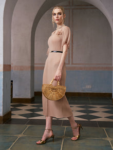 A woman in the Asymmetric Fringe Midi Dress with belt by Social Queen and gold heels stands indoors, holding a wooden handbag. She is in profile with her hair styled back, in a tiled room with arched doorways.