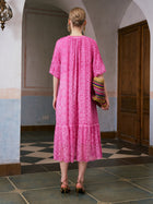A woman stands indoors facing away, wearing the Social Queen Geometric Jacquard Mulberry Silk Dress and heels, holding a colorful woven clutch. The room features tiled floors, arched walls, and wooden doors—an elegant setting perfect for resort wear.