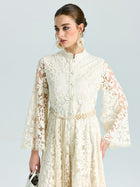A woman models the Embroidered Lace High-Neck Dress by Social Queen—featuring a long cream baroque floral design with bell sleeves and tied belt—styled with a white purse. Ideal as a special occasion dress thanks to its delicate lace-effect embroidery.