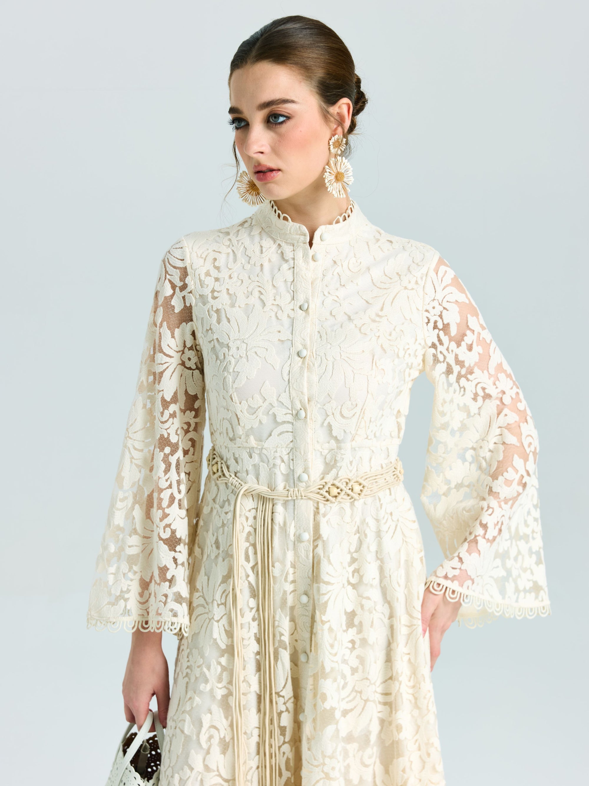 A woman models the Embroidered Lace High-Neck Dress by Social Queen—featuring a long cream baroque floral design with bell sleeves and tied belt—styled with a white purse. Ideal as a special occasion dress thanks to its delicate lace-effect embroidery.