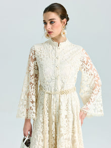 A woman models the Embroidered Lace High-Neck Dress by Social Queen—featuring a long cream baroque floral design with bell sleeves and tied belt—styled with a white purse. Ideal as a special occasion dress thanks to its delicate lace-effect embroidery.
