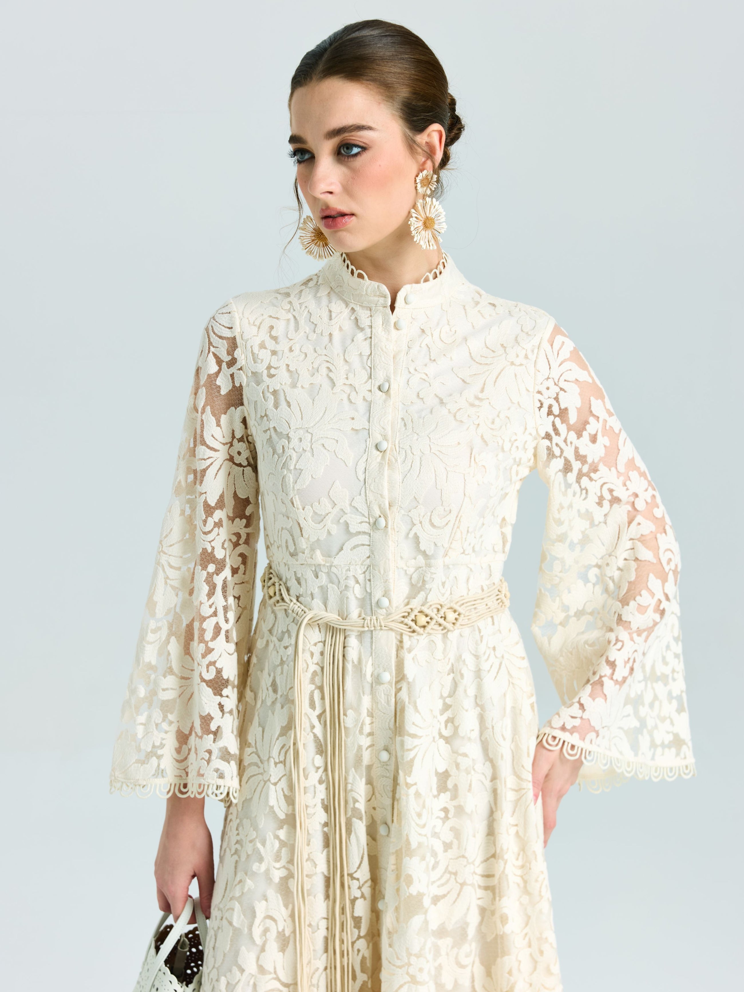 A woman models the Embroidered Lace High-Neck Dress by Social Queen—featuring a long cream baroque floral design with bell sleeves and tied belt—styled with a white purse. Ideal as a special occasion dress thanks to its delicate lace-effect embroidery.