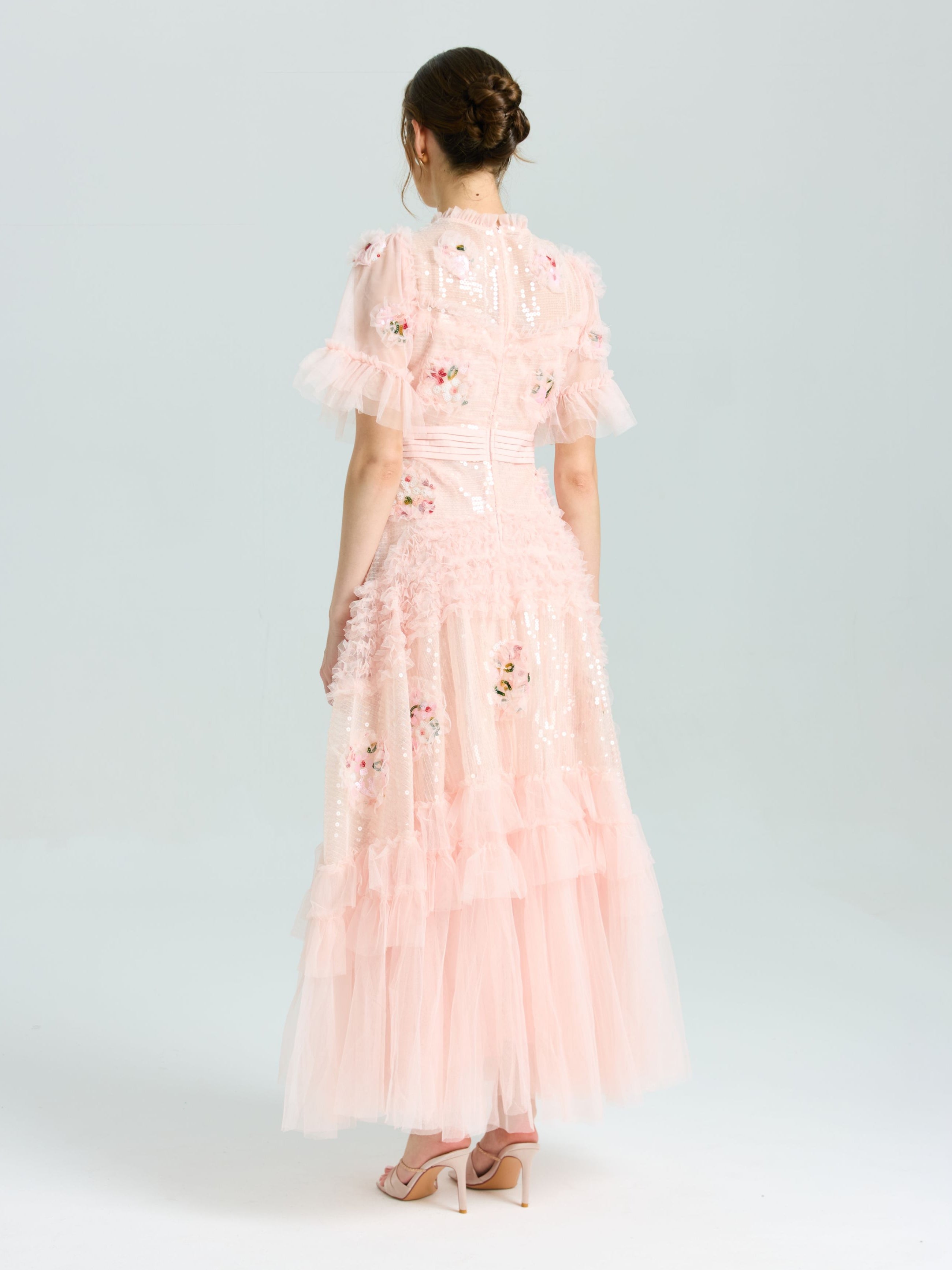 A woman faces away, wearing the Social Queen Embroidered Ruffle Tiered Tulle Dress in light pink with floral and sequin embroidery—an elegant choice for special occasions, styled with high heels against a plain light background.