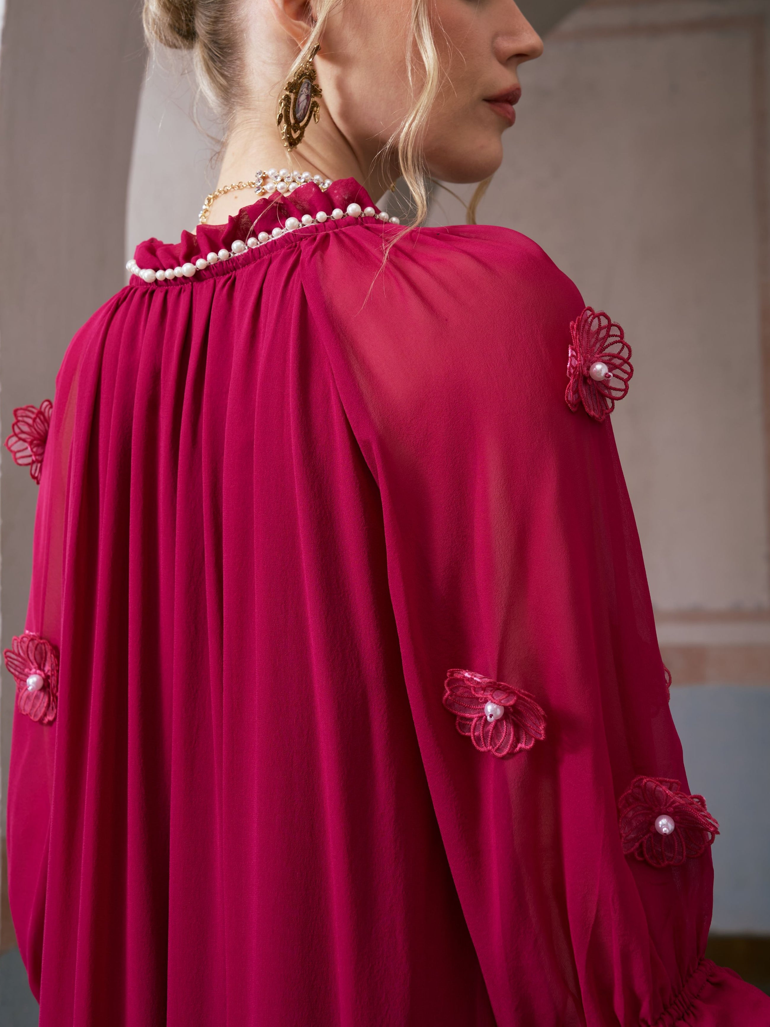 A woman wears the Social Queen 3D Floral Pearl Mulberry Silk Dress, featuring sheer sleeves, magenta 3D floral appliqué, and delicate pearl details on the collar. Her hair is styled up, and she accessorizes with gold earrings and a necklace.