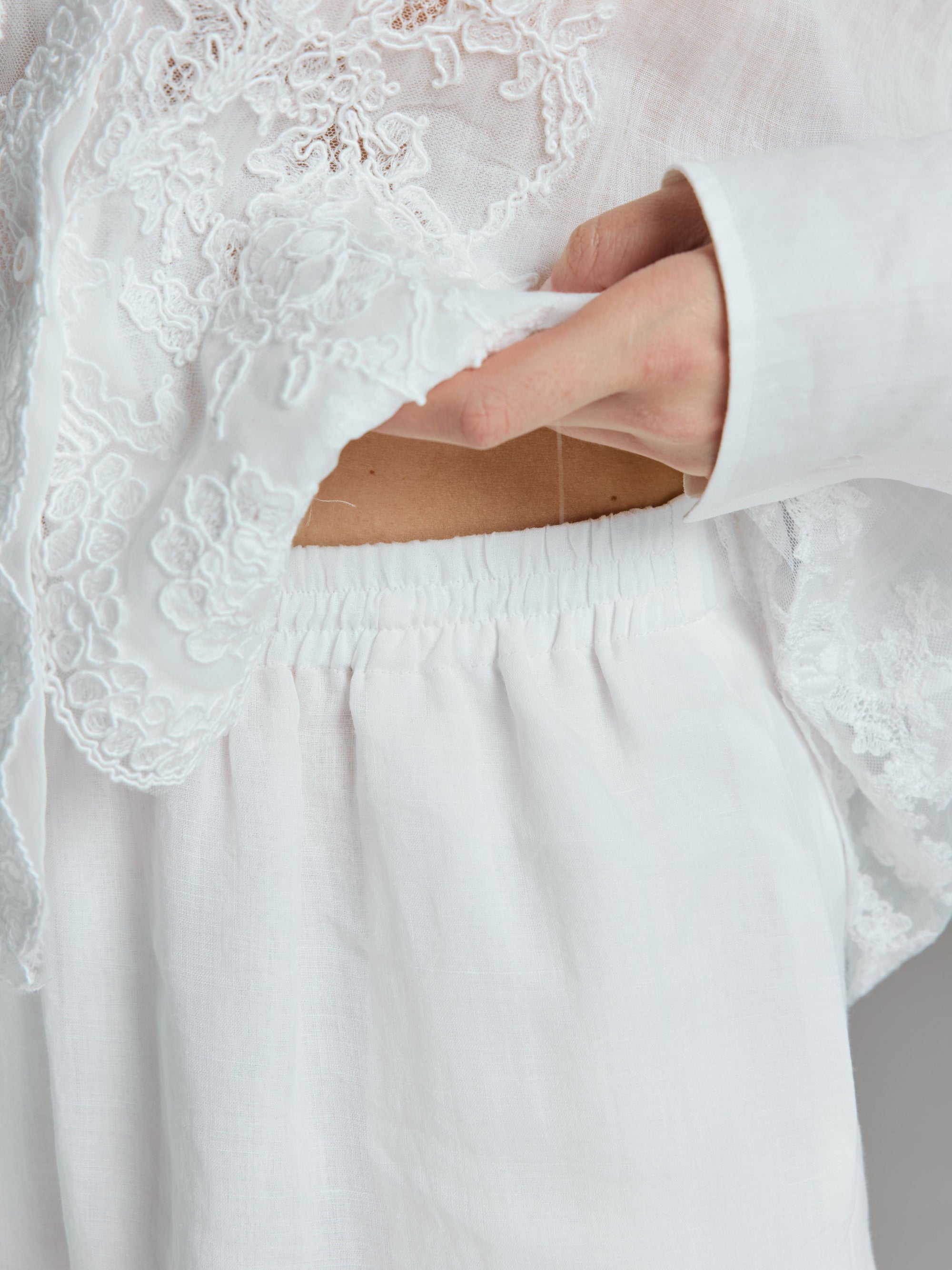 Wearing Social Queen’s Textured Straight-Leg Pants, a person in white lace and linen clothing lifts the hem of their top to reveal part of their bare midriff above the lightweight slub-textured pants.