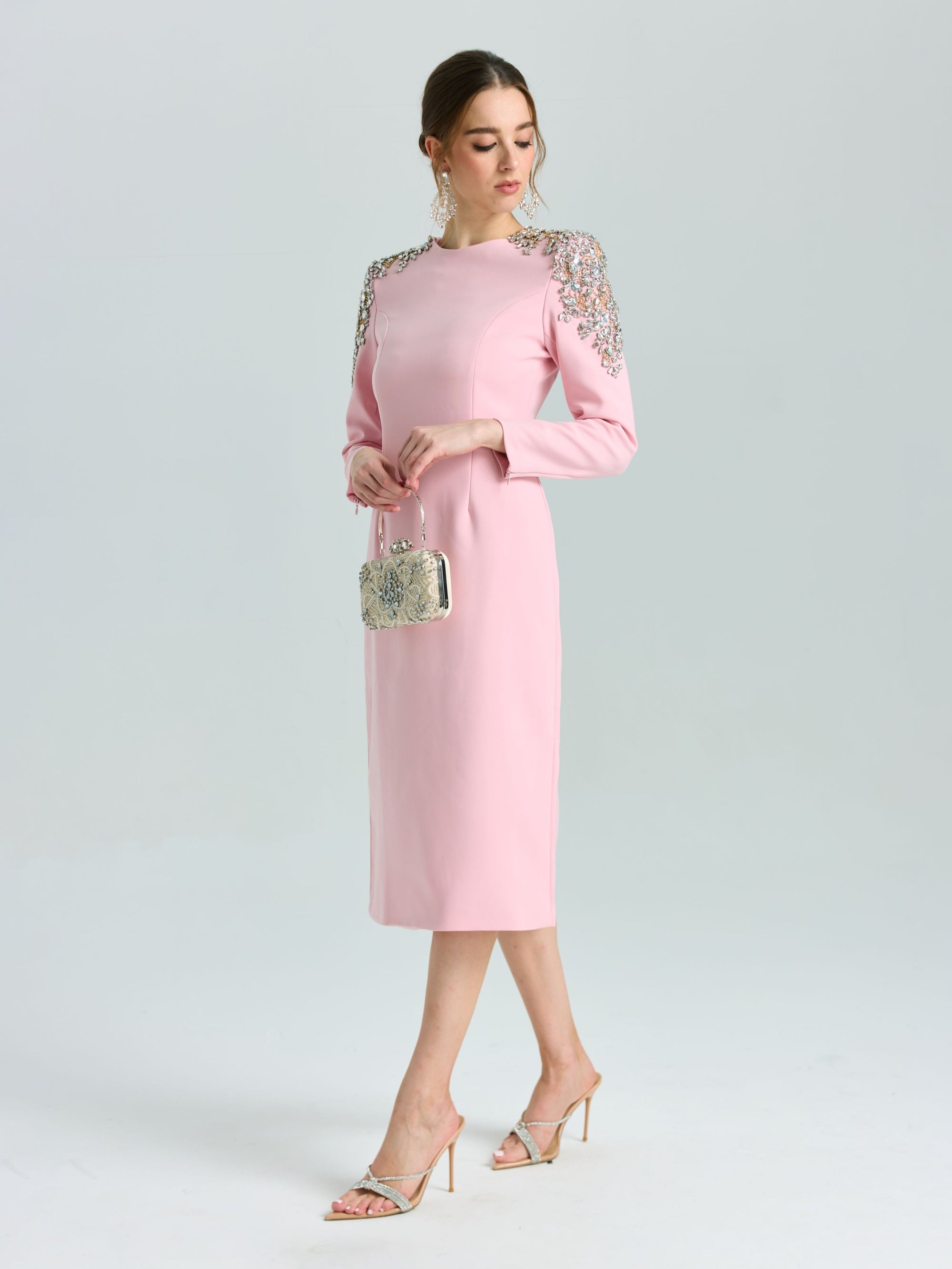 A woman wears the Social Queen Crystal Beaded Detail Midi Dress, featuring pink crepe fabric and crystal-embellished shoulders, styled with a metallic purse and heeled sandals—an elegant choice for evening events.