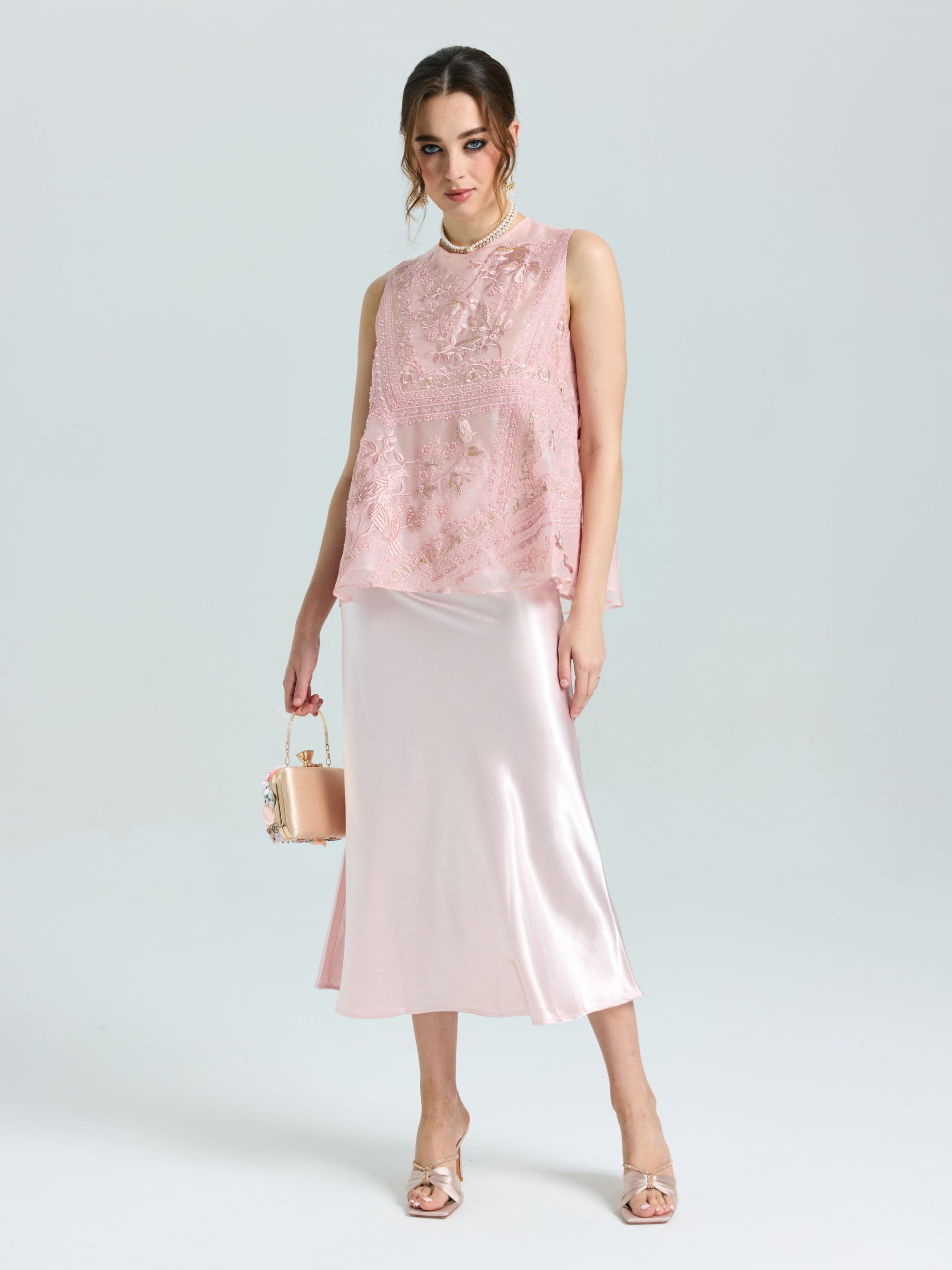 Woman wears the Social Queen Floral Bird Embroidered Silk Top—a sleeveless pink silk piece—paired with a shiny light pink midi skirt, open-toe heels, and a small pink handbag, posing against a plain light background.