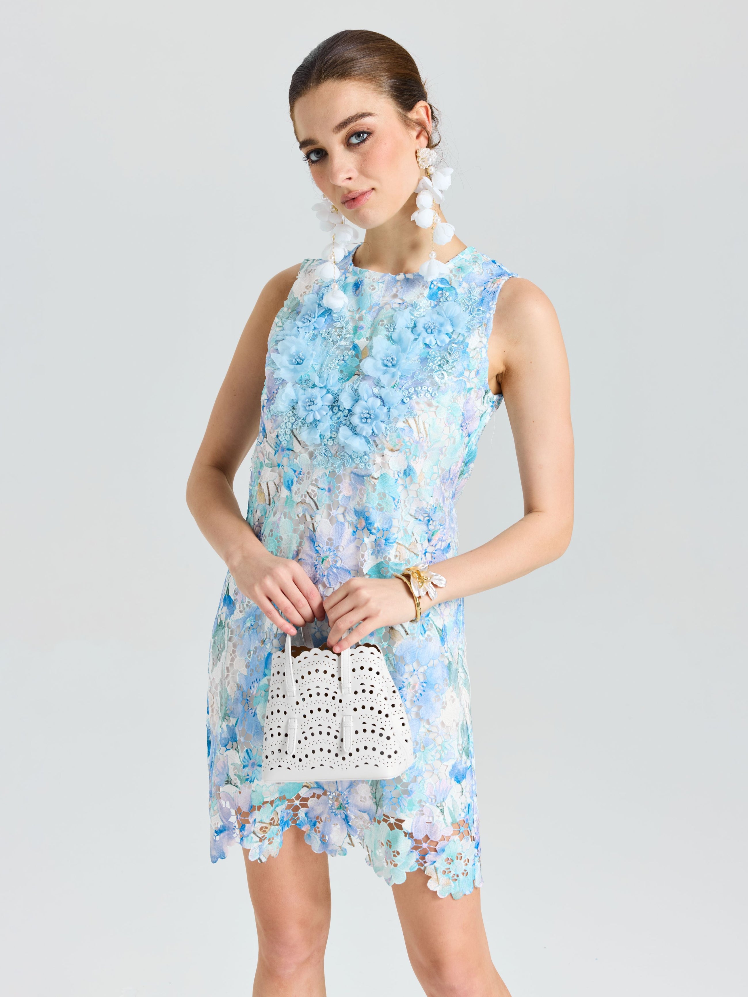A woman stands against a plain background in the Social Queen Embellished Floral Lace Mini Dress, styled with white statement earrings and a white perforated bag.