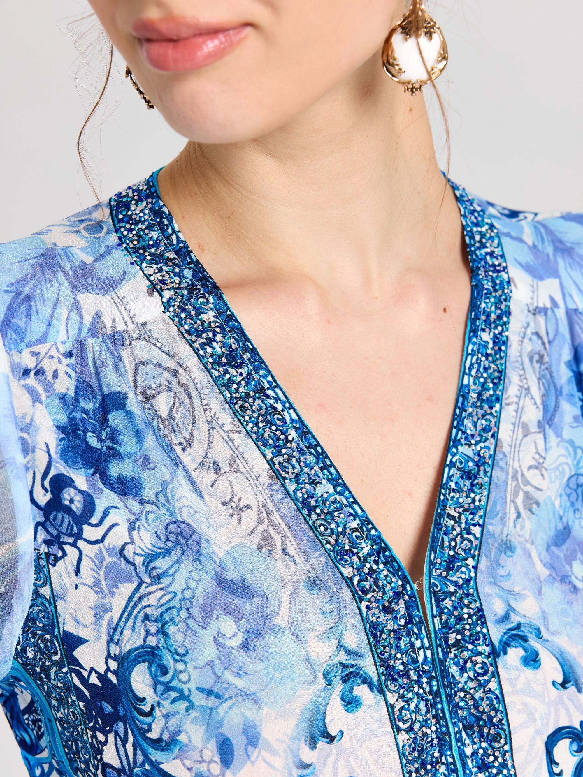 A woman wears the Baroque Placement Print Silk Dress by Social Queen, styled with gold earrings and light pink lipstick; her look echoes blue-and-white porcelain motifs, with her face partially visible.