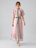 A woman stands against a plain background, wearing the Peacock Botanical Print Dress by Social Queen—a long pink A-line silhouette dress featuring floral patterns, subtle rhinestone accents, puff sleeves, a blue-green belt, and white heeled sandals.