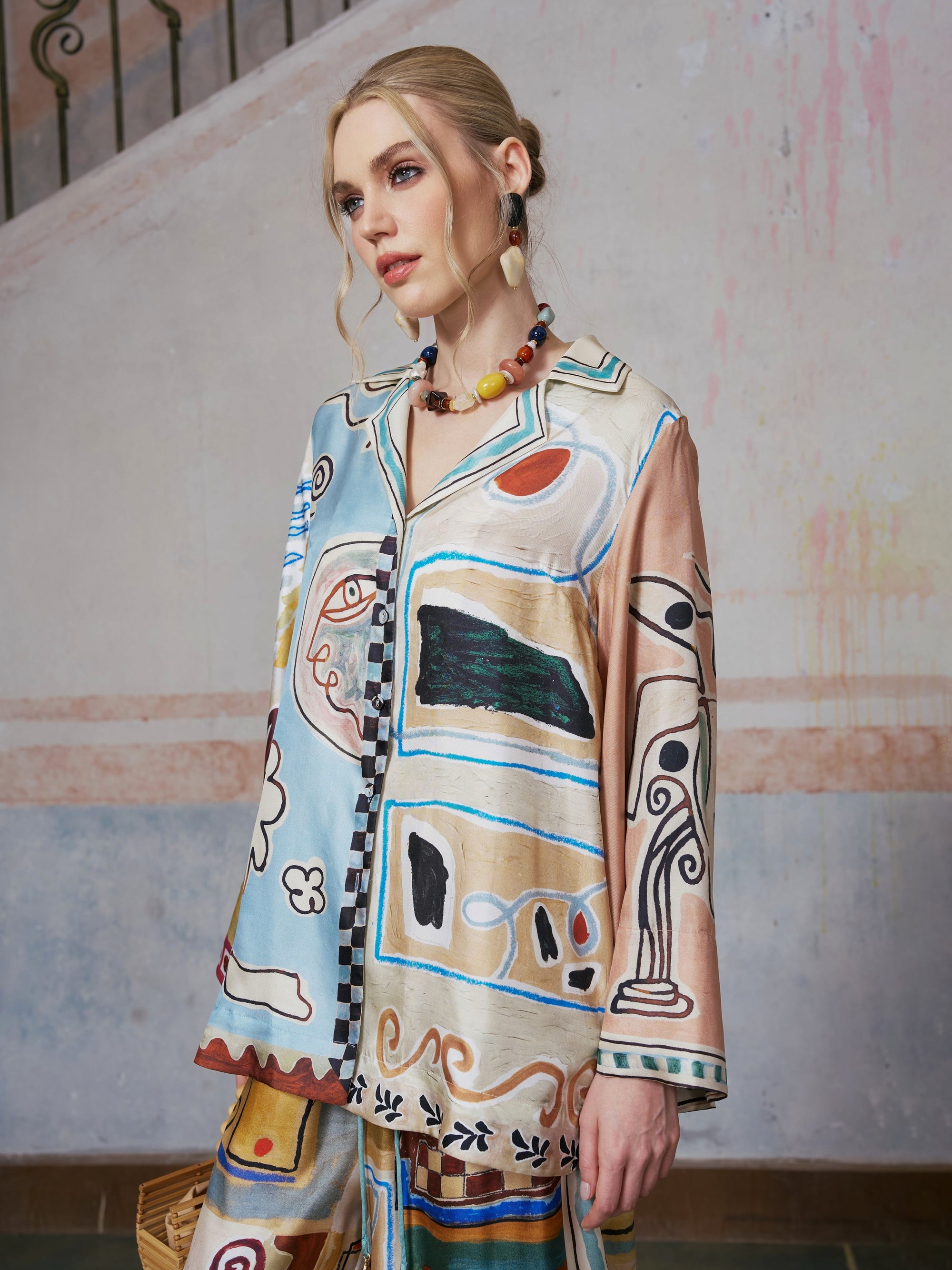 A woman with blonde hair wears the Social Queen Abstract Print Mulberry Silk Shirt and bold jewelry, standing indoors by a pastel wall and staircase—perfectly capturing chic resort wear style.