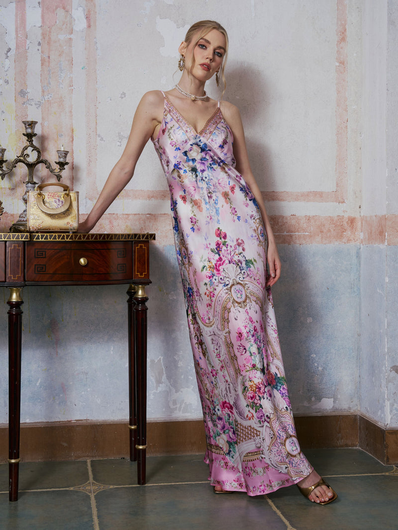 A woman wearing the Social Queen Baroque Print Mulberry Silk Dress stands beside a wooden table adorned with ornate decor, set against a textured, pastel-colored wall.