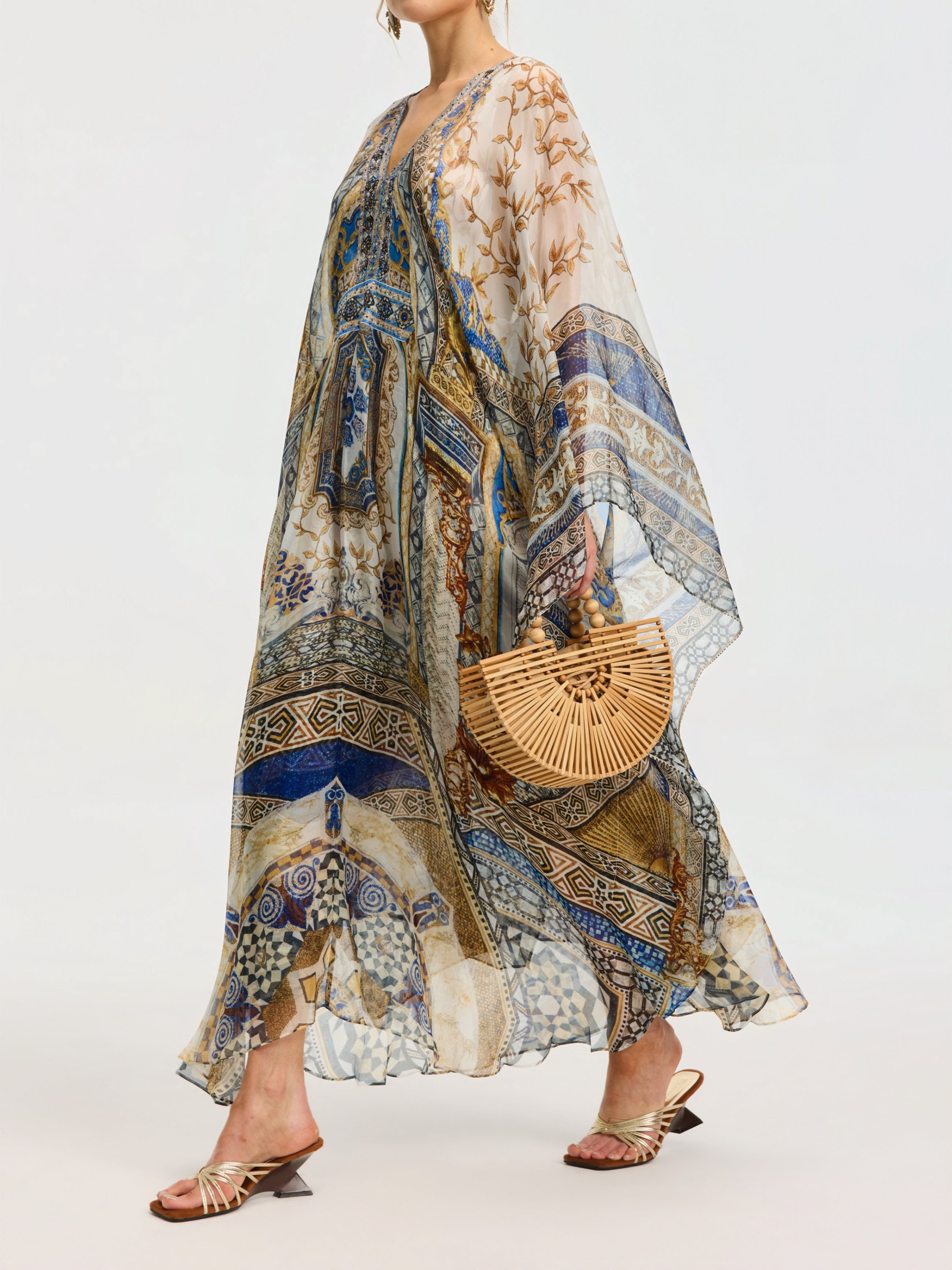A person wears the Social Queen Byzantine Print Silk Caftan Dress with blue and gold patterns, a crystal-embellished neckline, holding a wooden bag, and finishing the look with heeled sandals.