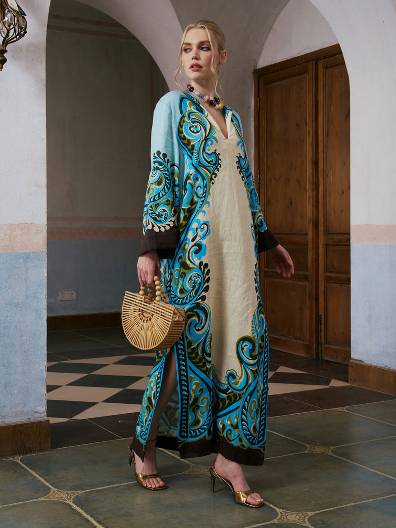 A woman wearing the Social Queen Paisley Print Linen Dress with a belt, paired with gold heels and holding a wooden handbag, stands indoors on a tiled floor near wooden doors.