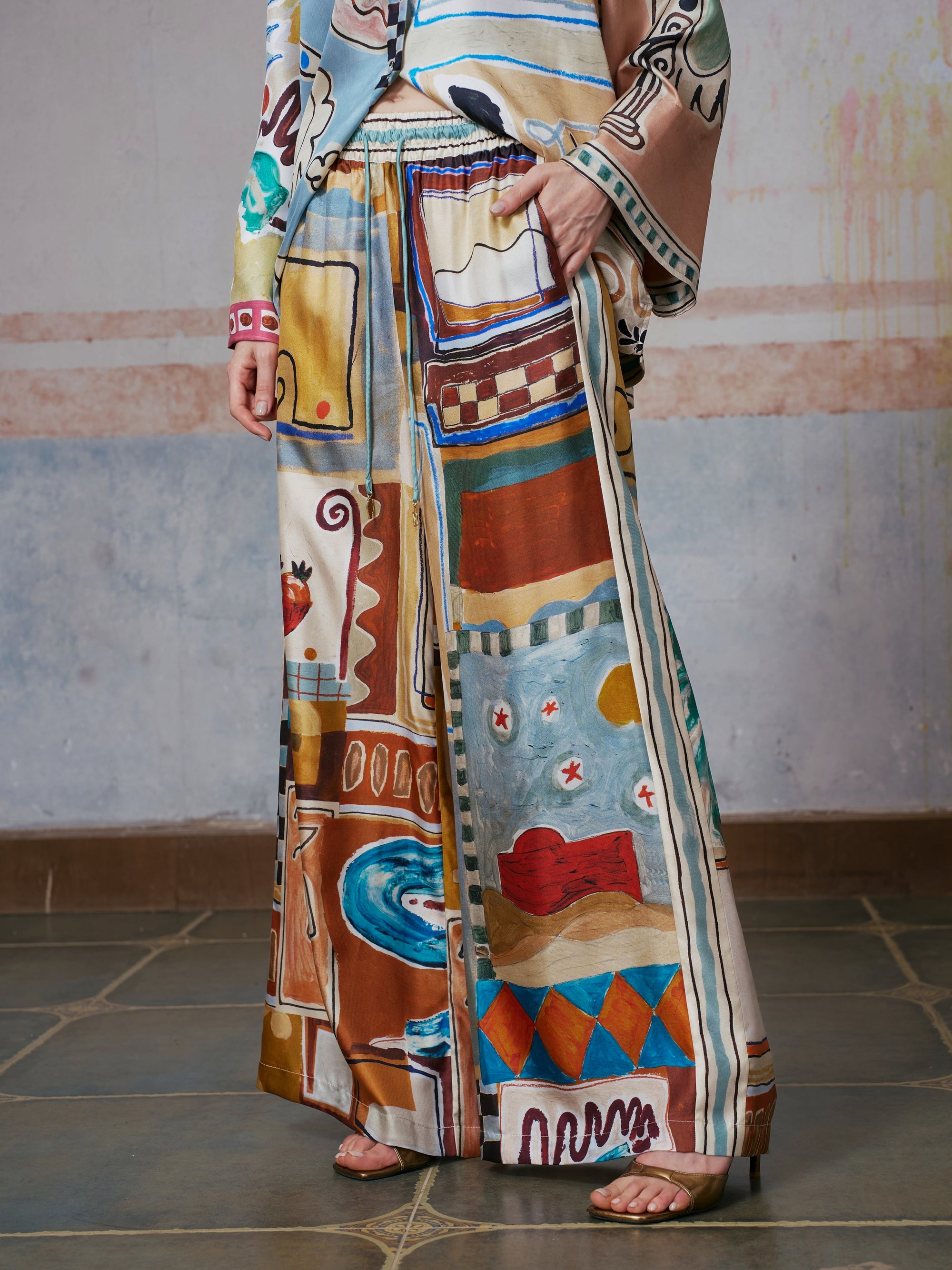 A person wearing Social Queen’s Abstract Print Mulberry Silk Pants with a matching top, featuring a colorful abstract design, stands on a tiled floor with one hand in the pants pocket. The face is out of frame.