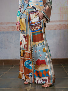 A person wearing Social Queen’s Abstract Print Mulberry Silk Pants with a matching top, featuring a colorful abstract design, stands on a tiled floor with one hand in the pants pocket. The face is out of frame.