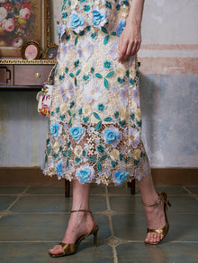 A person in gold heels wears the Social Queen 3D Floral Cutwork Lace Skirt with blue and white floral embroidery, holding a small bag while standing on a tiled floor.