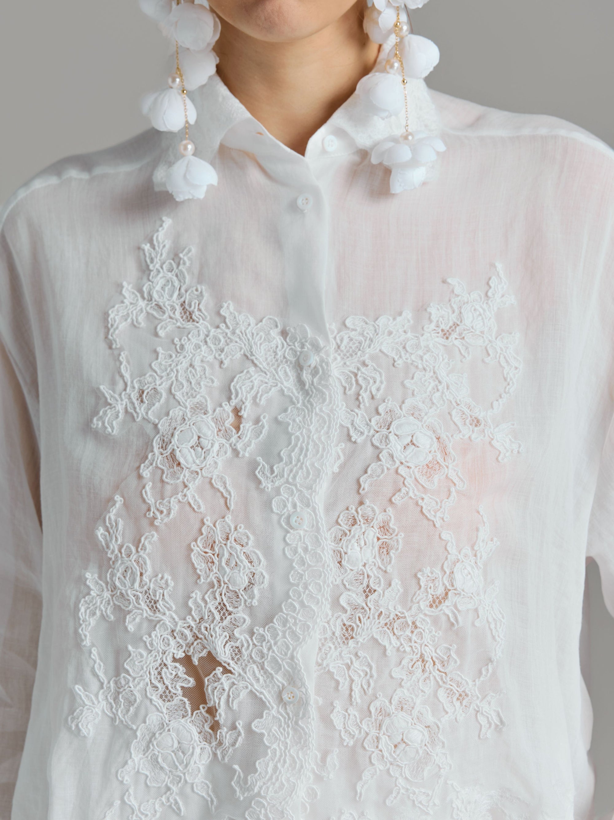 A person wearing the Social Queen 3D Lace Appliqué Shirt, featuring sheer white rose vine lace and floral lace embroidery, paired with large white floral earrings, shown from shoulders to chest.