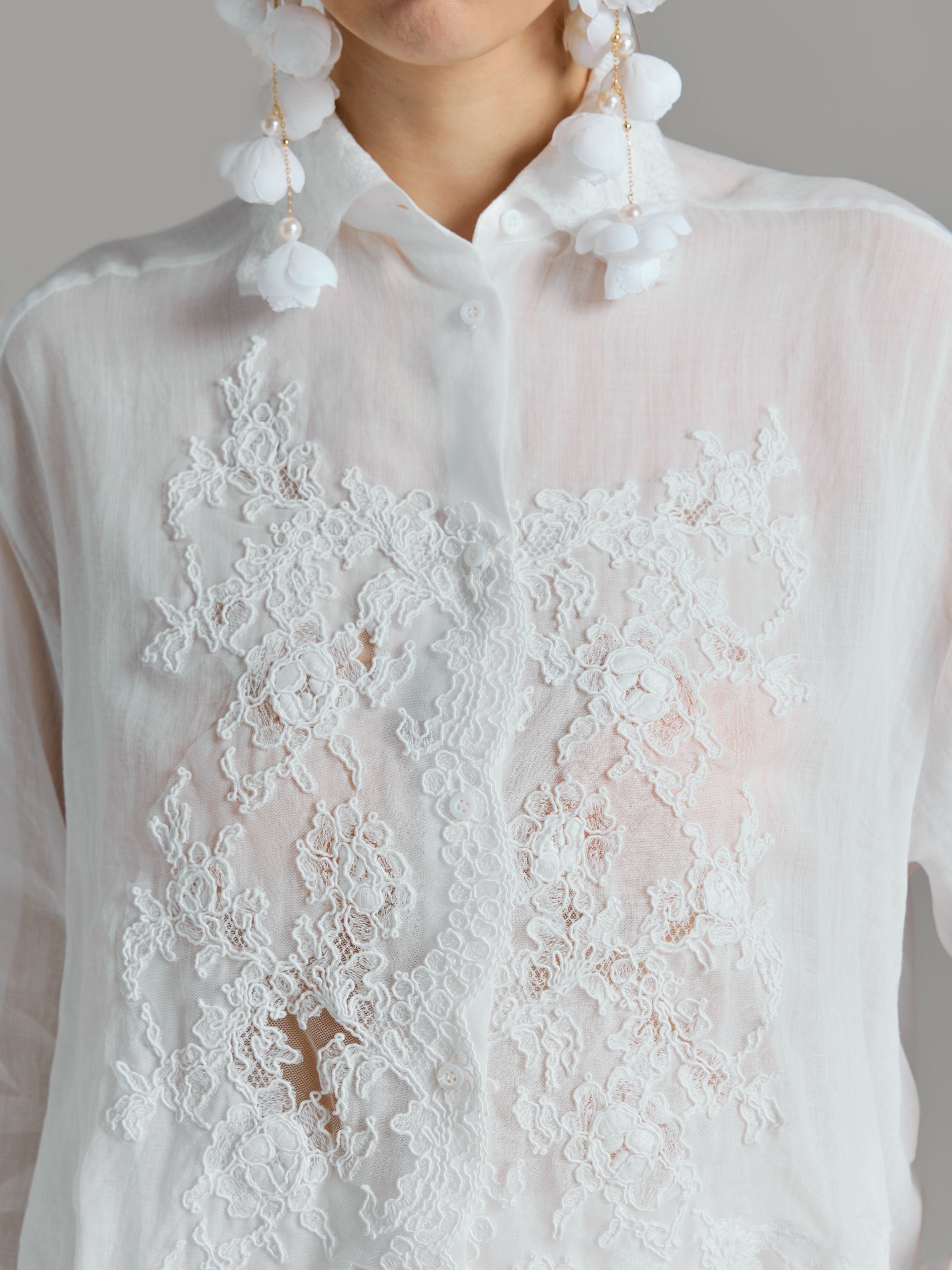 A person wearing the Social Queen 3D Lace Appliqué Shirt, featuring sheer white rose vine lace and floral lace embroidery, paired with large white floral earrings, shown from shoulders to chest.