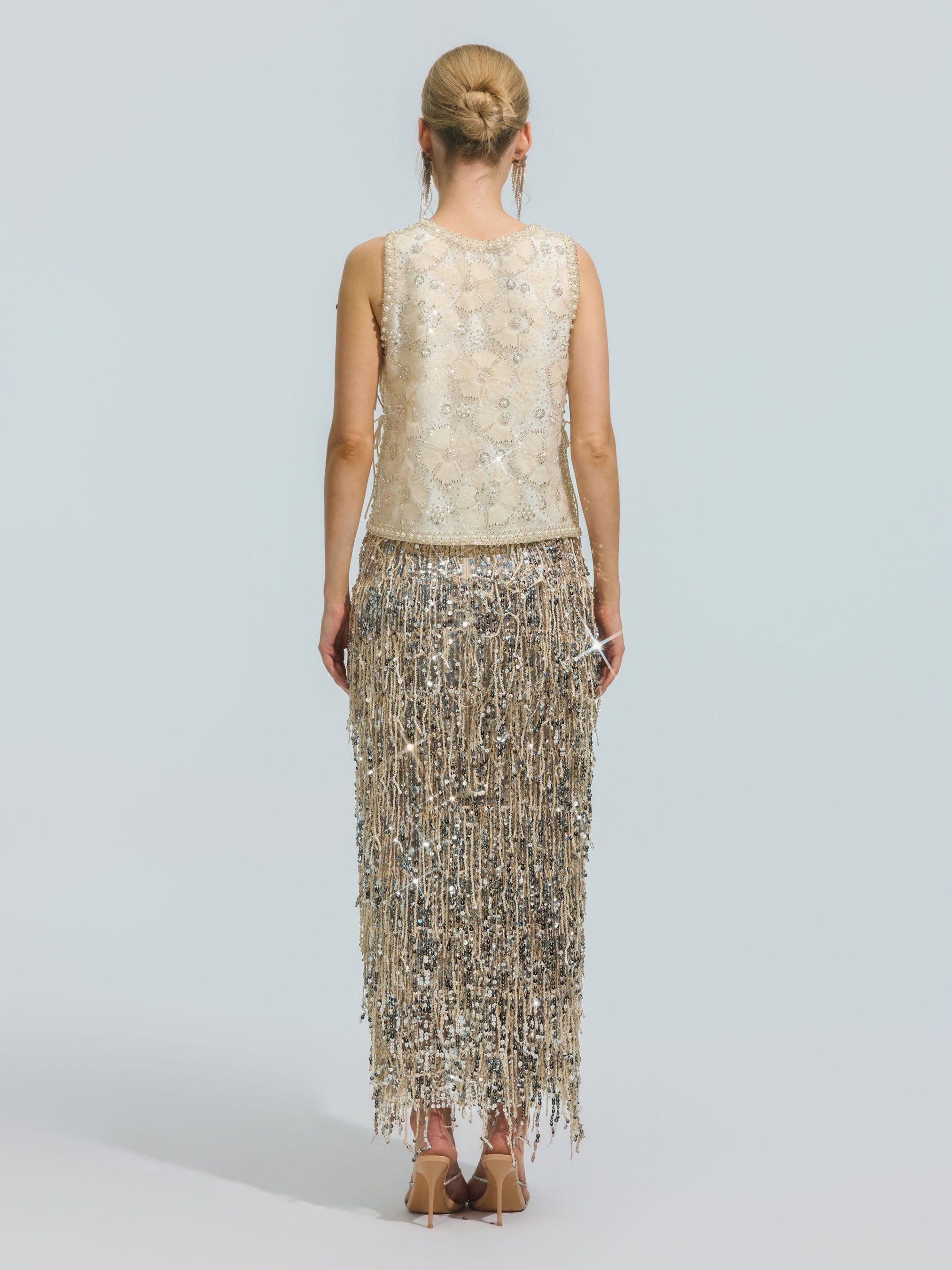 A woman stands facing away, wearing the Social Queen Floral Beaded Mesh Sleeveless Top paired with a long skirt featuring silver fringe, set against a plain light background.