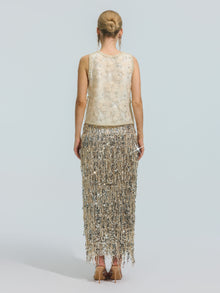 A woman stands facing away, wearing the Social Queen Floral Beaded Mesh Sleeveless Top paired with a long skirt featuring silver fringe, set against a plain light background.