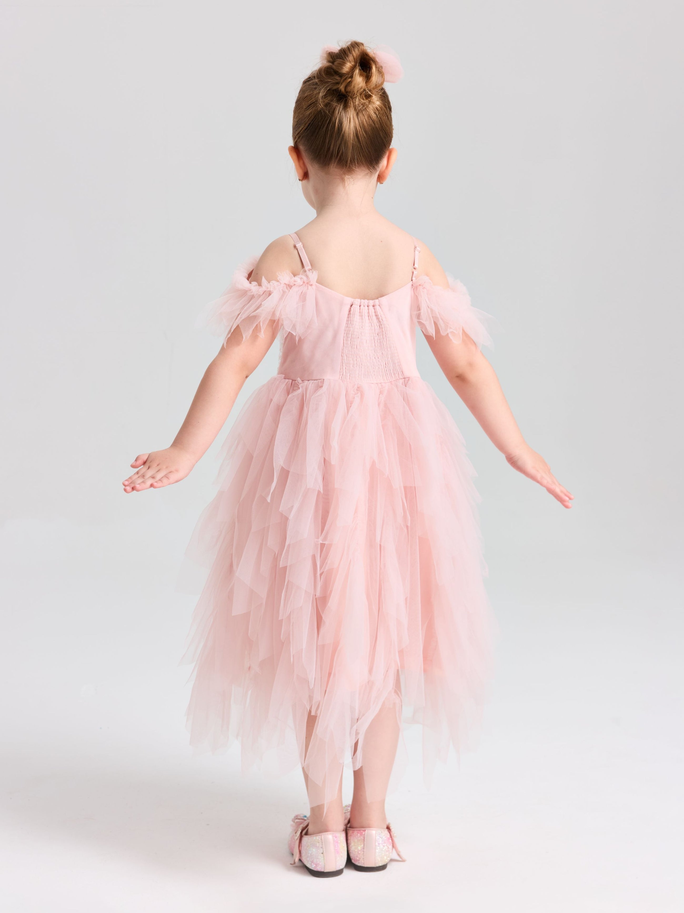 A young girl wears the Social Queen Girls Floral Sequin Tulle Dress, featuring an off-the-shoulder pink design, with matching shoes, standing arms outstretched and facing away against a plain light background.