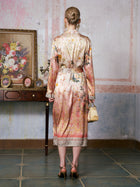 A woman with blond hair in a neat bun, dressed in the Social Queen Garden Print Mulberry Silk Shirt Dress paired with heels, stands facing a wall in a vintage room adorned with a painting, small table, and tiled floor. The baroque garden print of her designer resort wear infuses the timeless scene with refined elegance.
