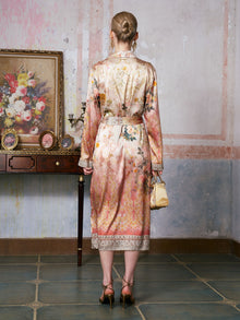 A woman with blond hair in a neat bun, dressed in the Social Queen Garden Print Mulberry Silk Shirt Dress paired with heels, stands facing a wall in a vintage room adorned with a painting, small table, and tiled floor. The baroque garden print of her designer resort wear infuses the timeless scene with refined elegance.