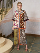 A woman stands on a tiled floor near stairs, wearing the Social Queen Patchwork Print Cotton Shirt Dress (belt included), paired with sandals and holding a brown handbag, with light-colored walls in the background.