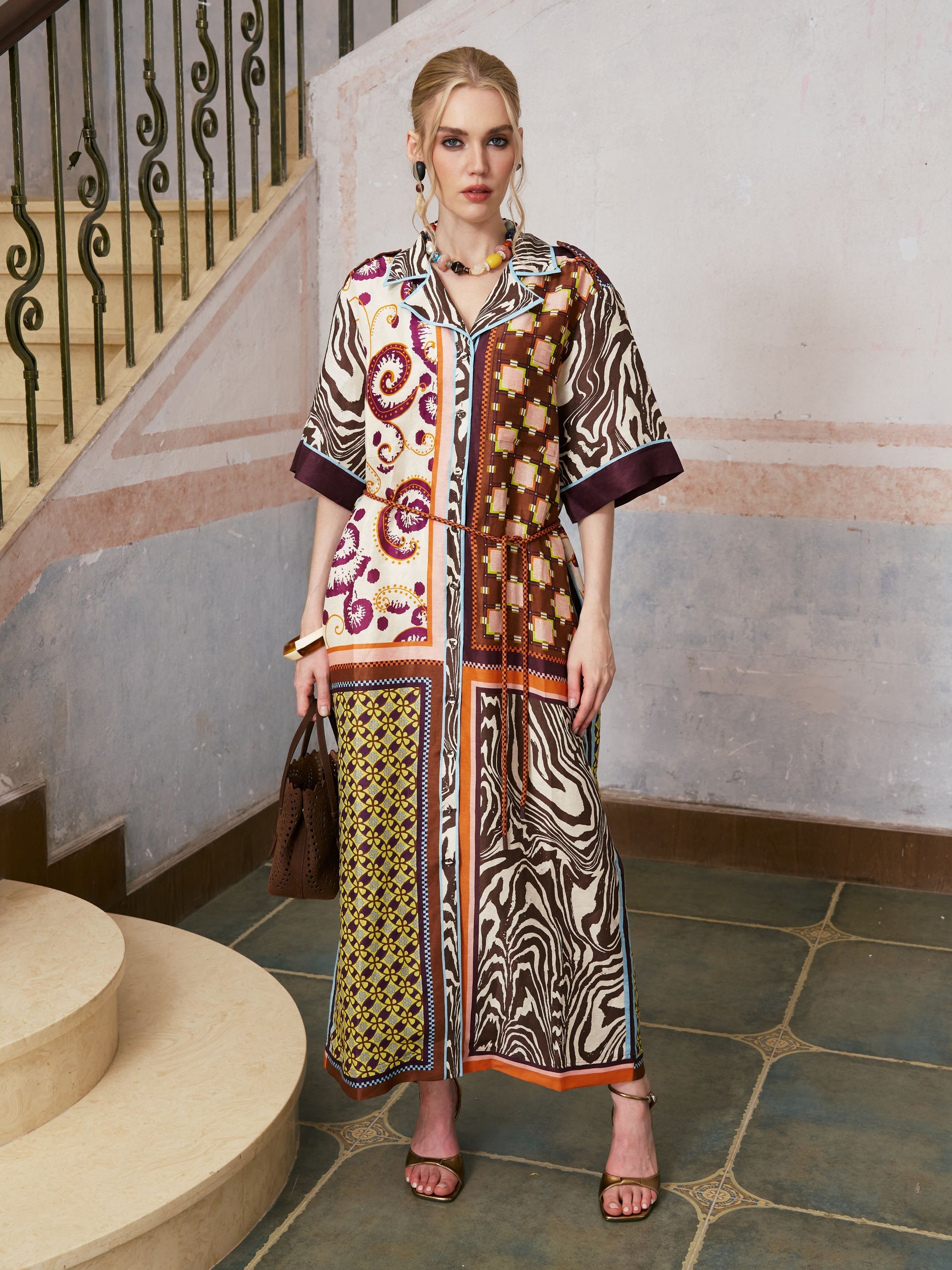 A woman stands on a tiled floor near stairs, wearing the Social Queen Patchwork Print Cotton Shirt Dress (belt included), paired with sandals and holding a brown handbag, with light-colored walls in the background.