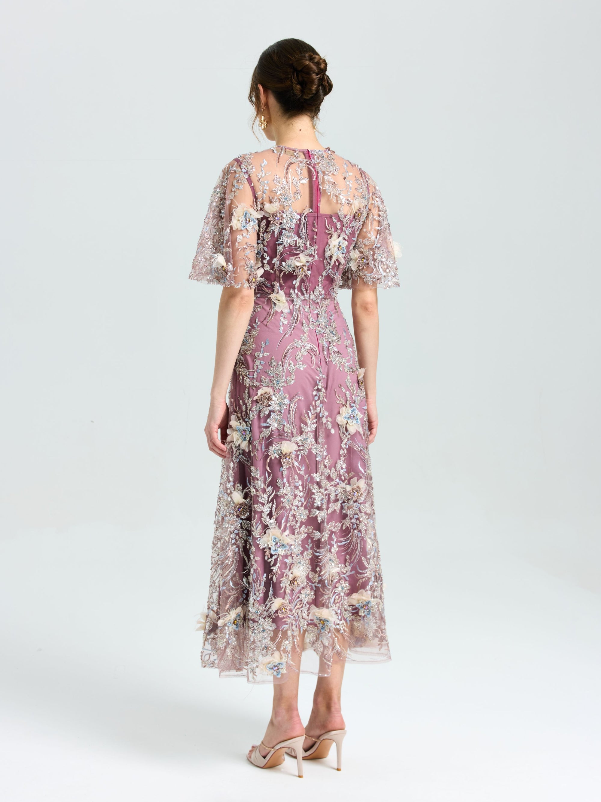 A woman faces away in the Social Queen Floral Beaded A-Line Dress, featuring lavender floral embroidery and sheer short sleeves, paired with nude heels against a plain light background.