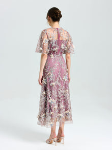 A woman faces away in the Social Queen Floral Beaded A-Line Dress, featuring lavender floral embroidery and sheer short sleeves, paired with nude heels against a plain light background.