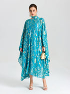 A woman models the Social Queen Gold Botanical Jacquard Silk Dress, featuring a beaded neckline. She pairs it with a gold handbag and open-toed heels, posing against a plain background.