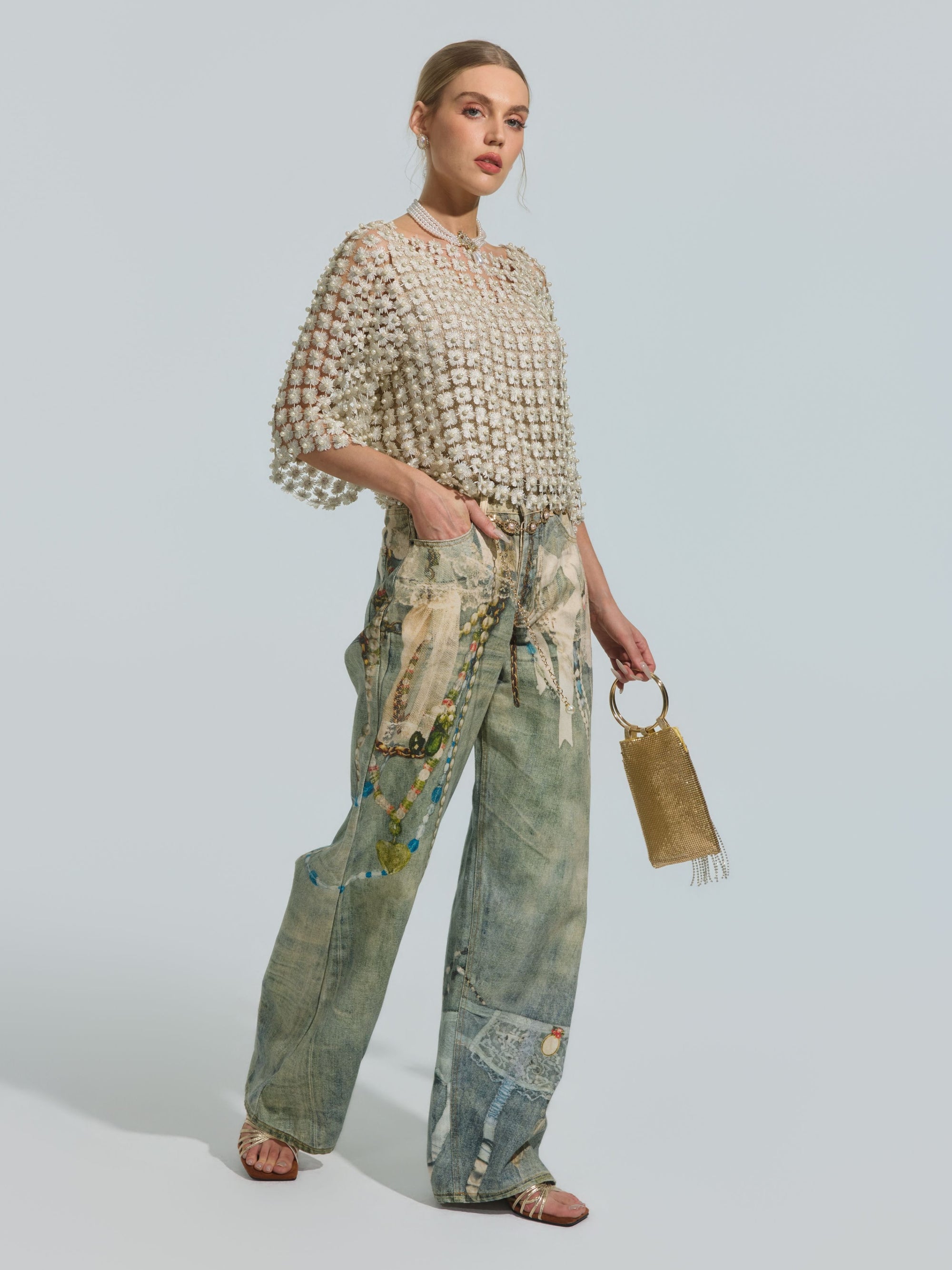 A woman stands wearing the Pearl Daisy Cutwork Top by Social Queen (camisole not included), paired with wide-leg patchwork jeans, sandals, and a gold handbag.