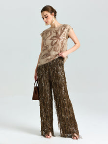 A woman poses in the Social Queen Floral Sequin Embroidered Silk Top with a floral pattern and brown fringed pants, holding a small brown handbag against a plain light background.