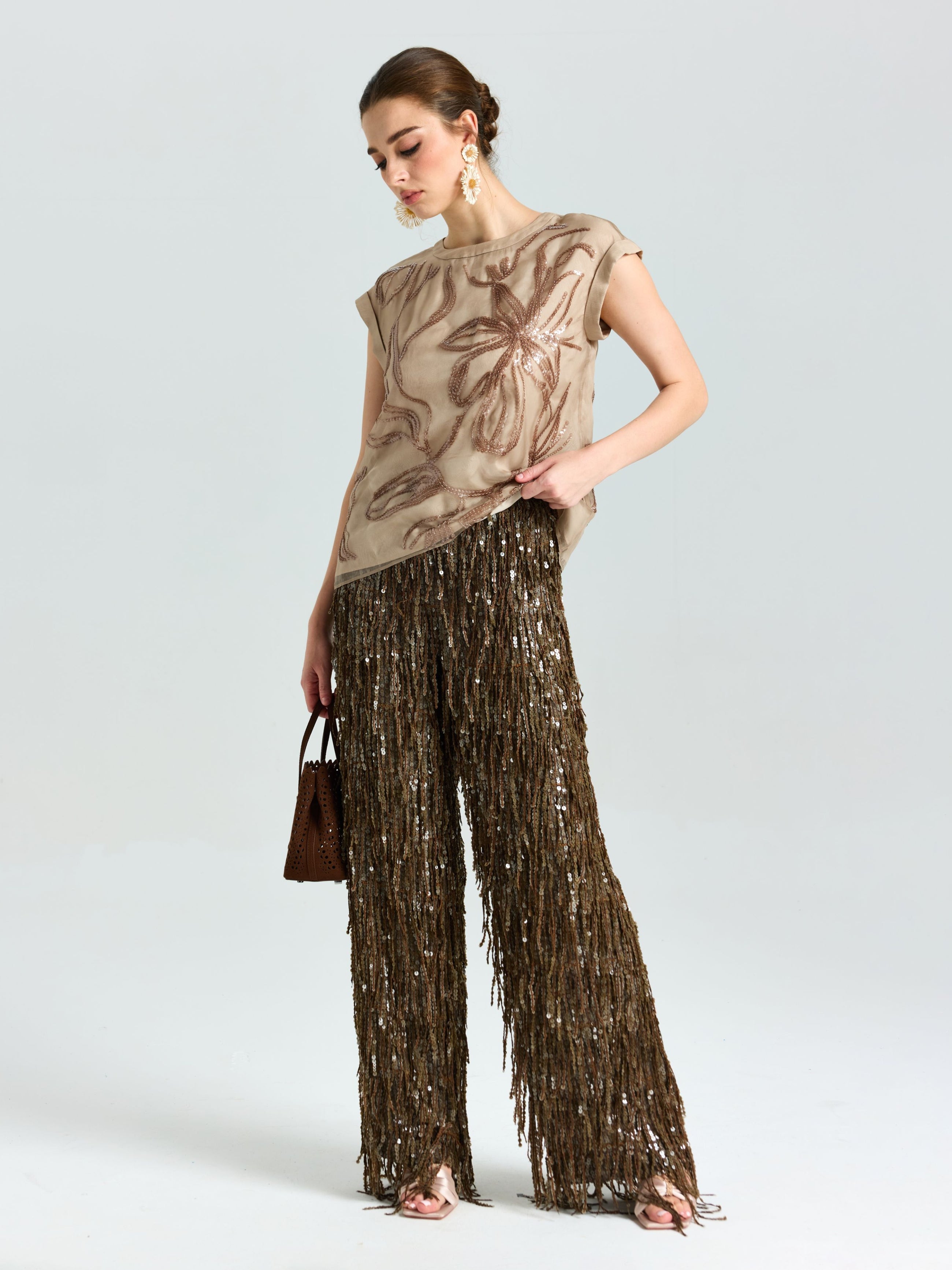 A woman poses in the Social Queen Floral Sequin Embroidered Silk Top with a floral pattern and brown fringed pants, holding a small brown handbag against a plain light background.