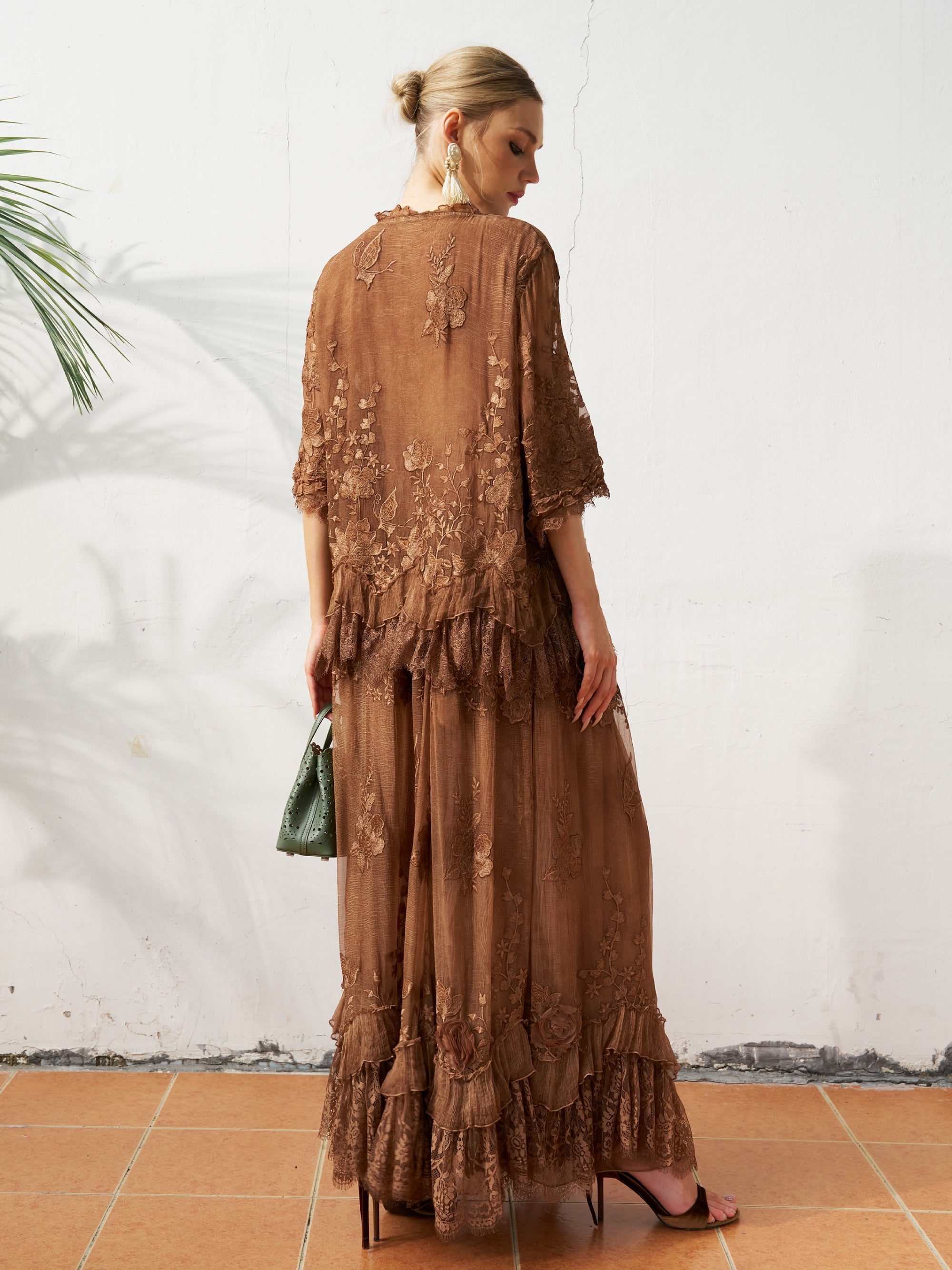 A woman stands outdoors on a tiled floor by a white wall and palm leaf, wearing the Social Queen 3D Floral Embroidered 100% Silk Ruffle Skirt. She faces away and holds a green handbag.
