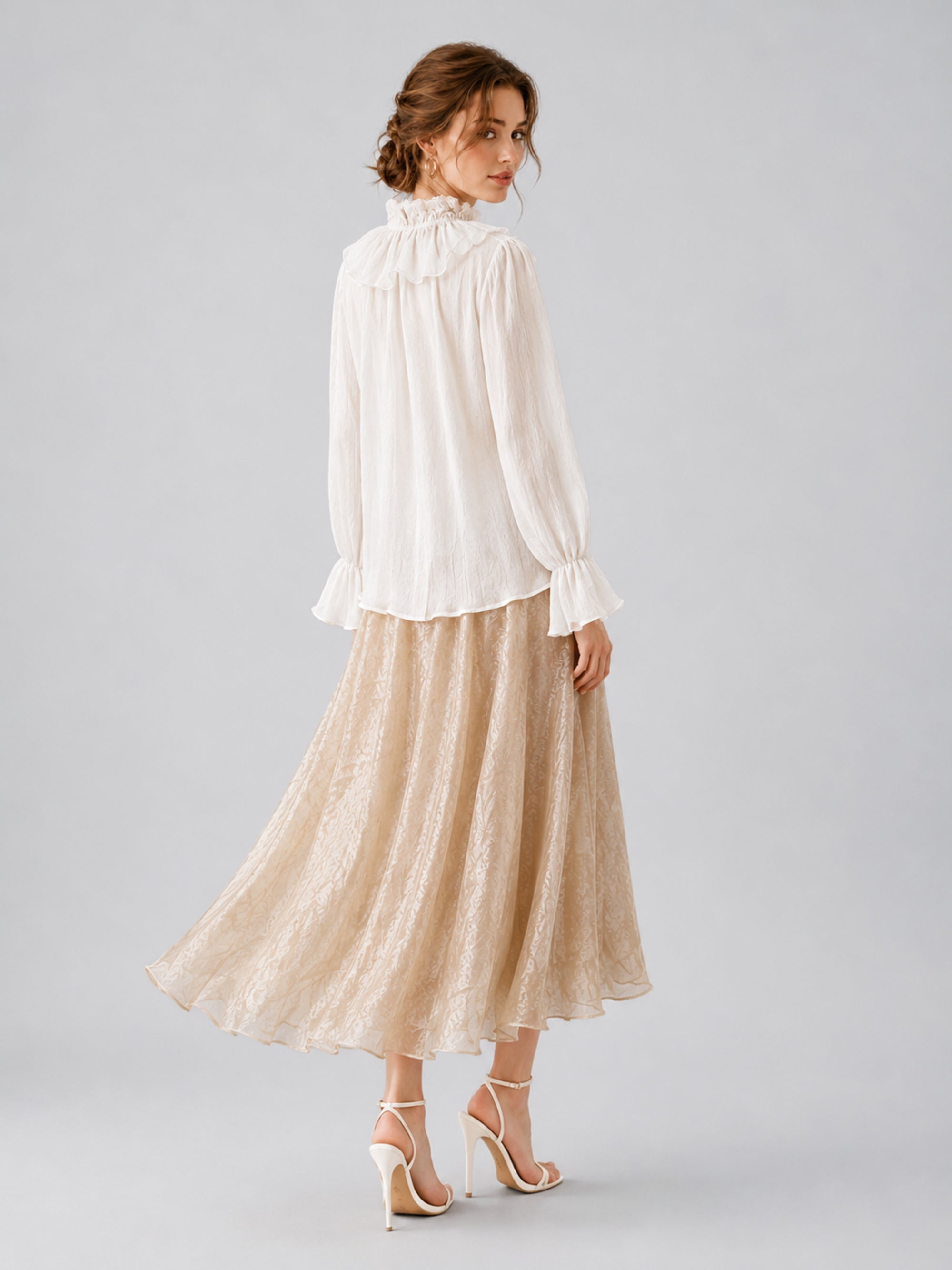 A woman stands looking over her shoulder, wearing a white long-sleeve blouse, the Social Queen Jacquard Chiffon Midi Skirt in beige with delicate lace detailing, and white high-heeled sandals against a plain gray background.